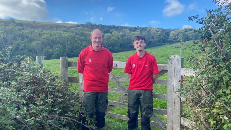 Two rangers in red standing in front of a countryside gate.